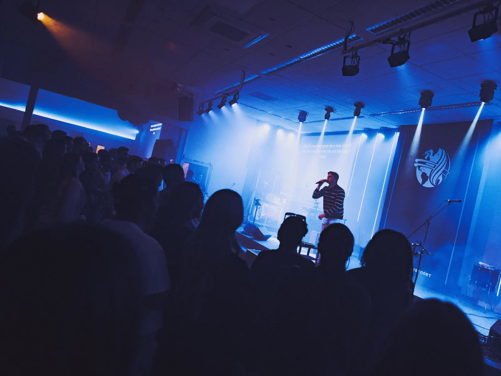 Volle zaal met jongeren in blauw licht, terwijl een zanger op het podium de aanbidding leidt